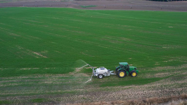 A green tractor towing a Tow and Fert Multi 2800 sprayer across a green pasture, applying liquid fertiliser through a spray boom.