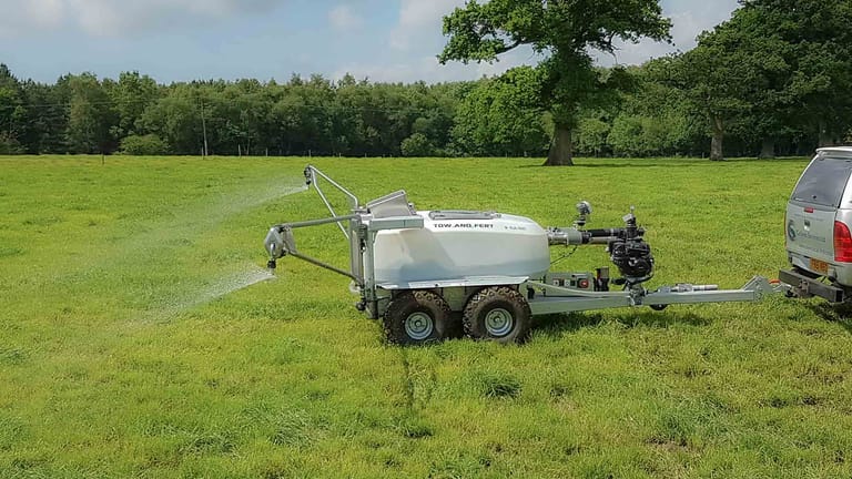 A Tow and Fert tow-behind sprayer attached to a utility vehicle, applying liquid fertiliser across a green pasture using its spray boom.