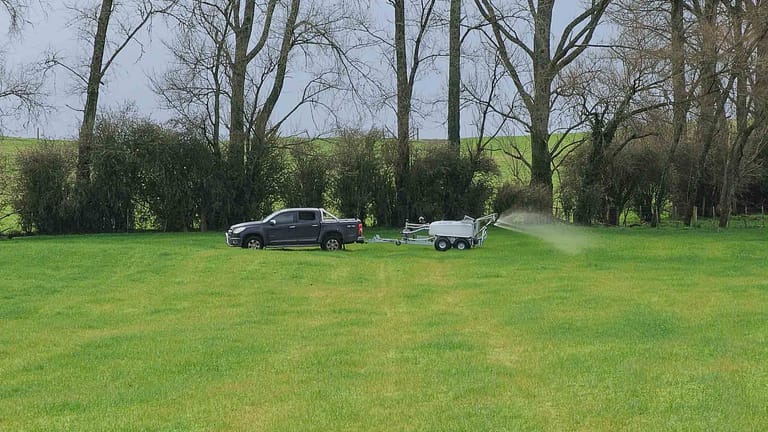 A grey pickup truck towing a Tow and Fert Multi 1000 sprayer across a green pasture, applying liquid fertiliser through an extended spray boom.