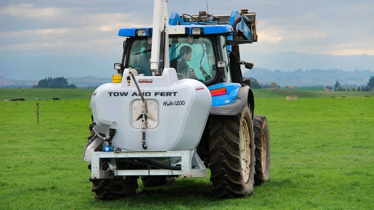 Rear view of a Tow and Fert Multi 1200 sprayer attached to a blue tractor, showing the sprayer tank, hoses and pump.