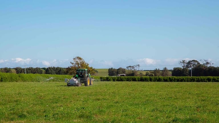 A green tractor towing a Tow and Fert Multi 1000 sprayer across a green pasture, applying liquid fertiliser through extended spray booms under a clear blue sky.